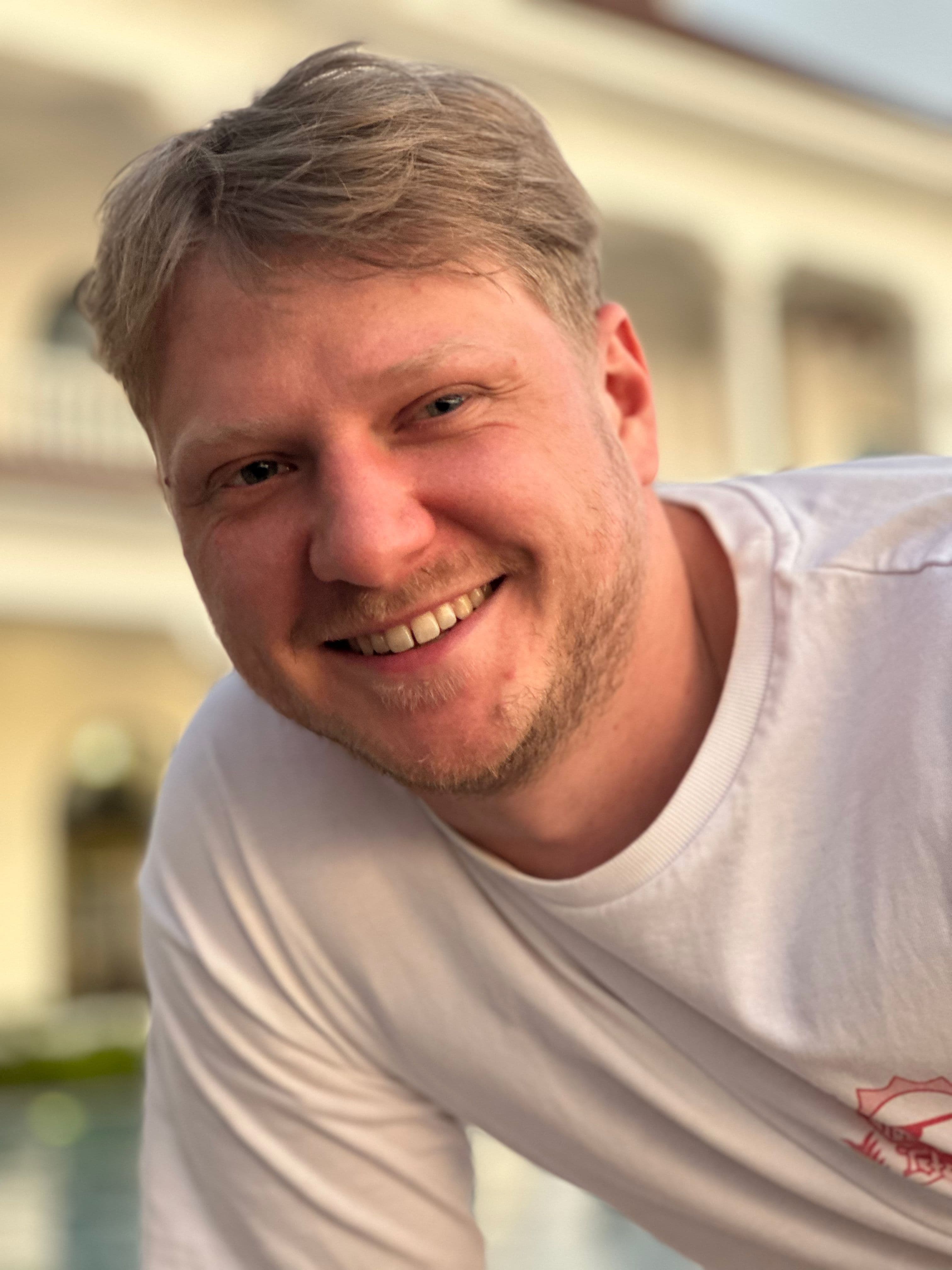 A smiling man with blond hair and a light beard wearing a white t-shirt.